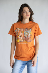 A woman modeling an orange graphic t-shirt with a 'Fall' theme featuring cowboy boots, pumpkins, and the text 'simply southern.' paired with light blue jeans, against a plain light gray background.