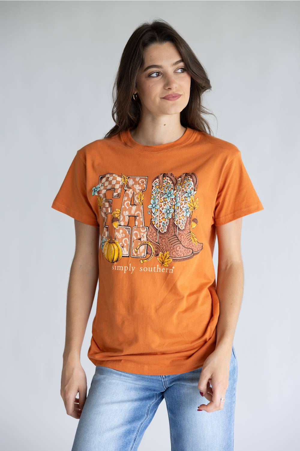 A woman modeling an orange graphic t-shirt with a 'Fall' theme featuring cowboy boots, pumpkins, and the text 'simply southern.' paired with light blue jeans, against a plain light gray background.
