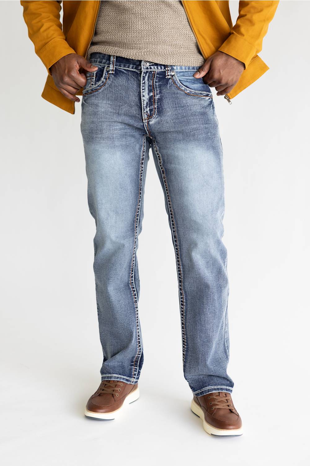 A person wearing light-wash blue denim jeans, a beige knit sweater, a mustard yellow jacket, and brown shoes, standing against a plain light background.