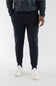 A person wearing black jogger sweatpants with a drawstring waist, paired with a dark patterned long-sleeve top and white sneakers, shown from the chest down on a light gray studio background.