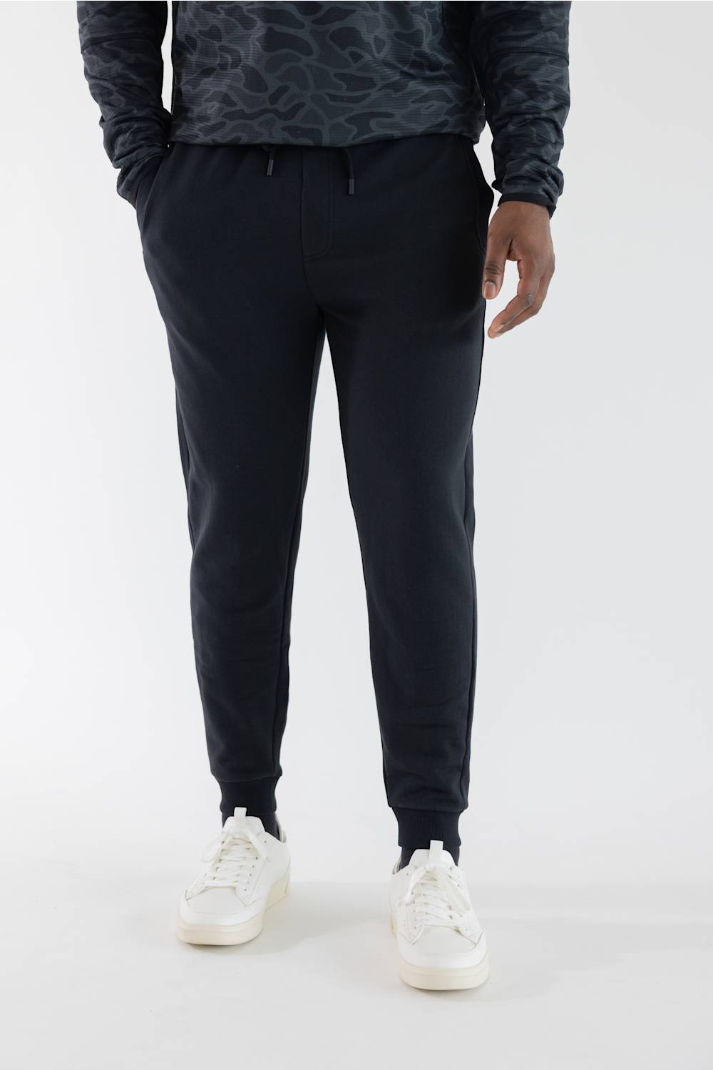 A person wearing black jogger sweatpants with a drawstring waist, paired with a dark patterned long-sleeve top and white sneakers, shown from the chest down on a light gray studio background.