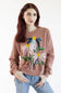 A woman with red hair wearing a dusty rose crewneck sweatshirt with a large floral applique design on the front, paired with blue jeans, against a plain white background.