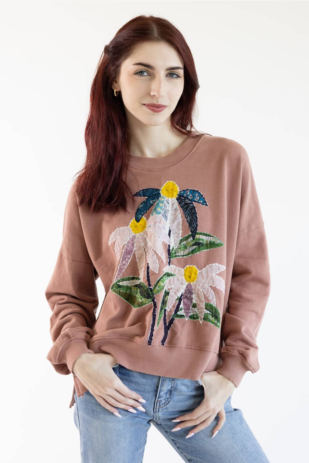 A woman with red hair wearing a dusty rose crewneck sweatshirt with a large floral applique design on the front, paired with blue jeans, against a plain white background.