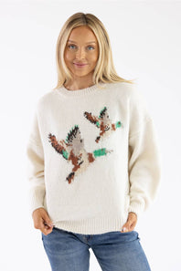 A woman wearing a cream-colored knit sweater with a large brown and green bird motif across the chest, paired with blue jeans, against a plain white background.