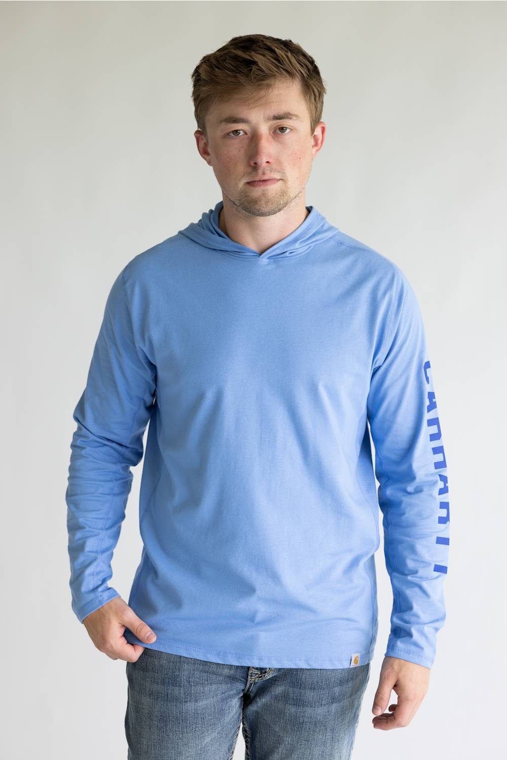 A man wearing a light blue long-sleeve hooded shirt with 'Canyon' printed vertically in blue on the right sleeve, paired with blue jeans, against a plain light gray background.