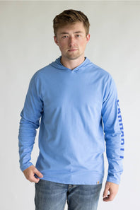 A man wearing a light blue long-sleeve hooded shirt with 'Canyon' printed vertically in blue on the right sleeve, paired with blue jeans, against a plain light gray background.