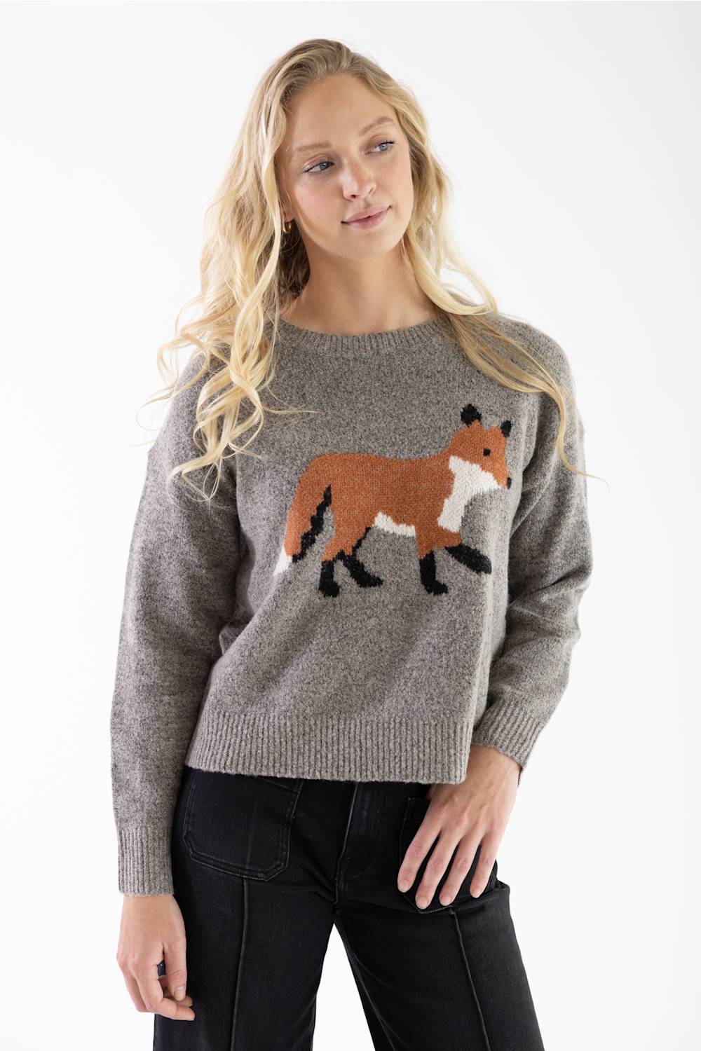 A woman wearing a heather gray knit sweater with a knitted fox motif in brown, white, and black, paired with black pants, against a plain white studio background.