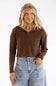 A woman wearing a brown cable-knit cropped zip-up sweater and light-wash blue jeans, standing against a plain white studio background.