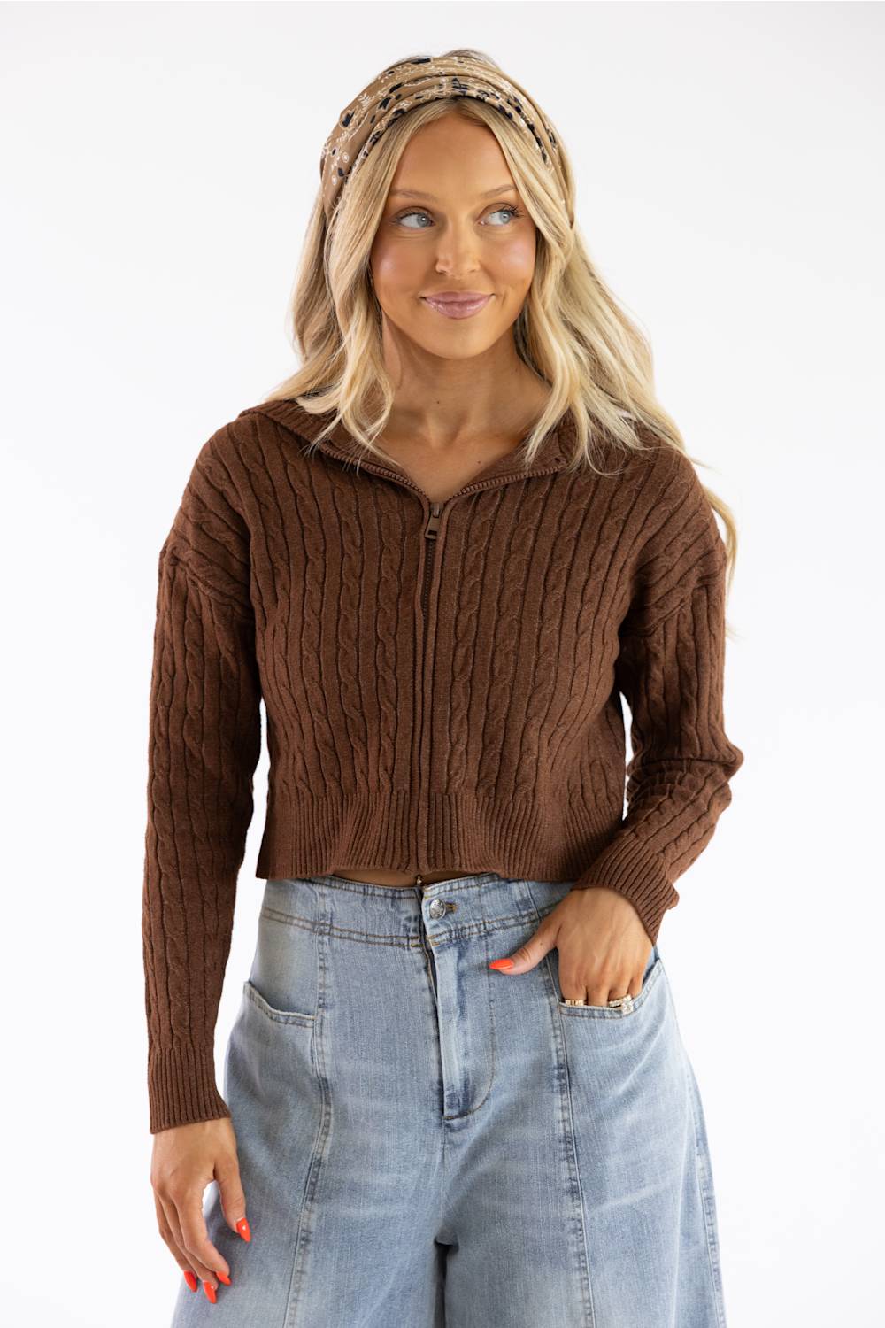 A woman wearing a brown cable-knit cropped zip-up sweater and light-wash blue jeans, standing against a plain white studio background.