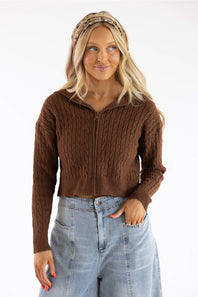 A woman wearing a brown cable-knit cropped zip-up sweater and light-wash blue jeans, standing against a plain white studio background.