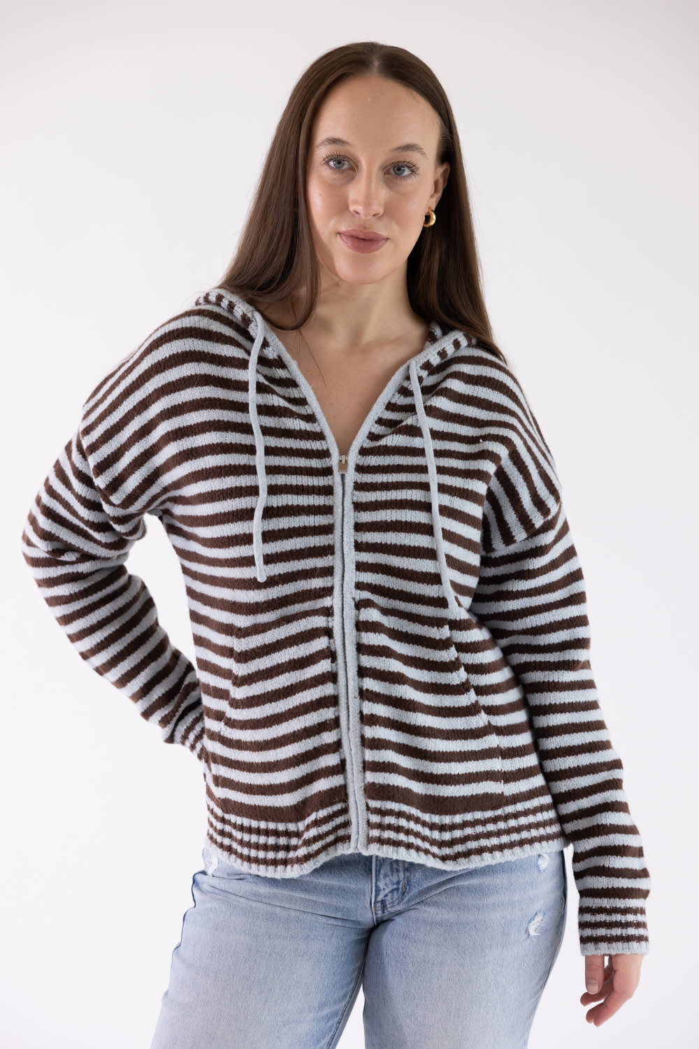 A woman modeling a brown and white horizontally striped zip-up hoodie with white drawstrings, paired with light blue jeans, against a plain white studio background.