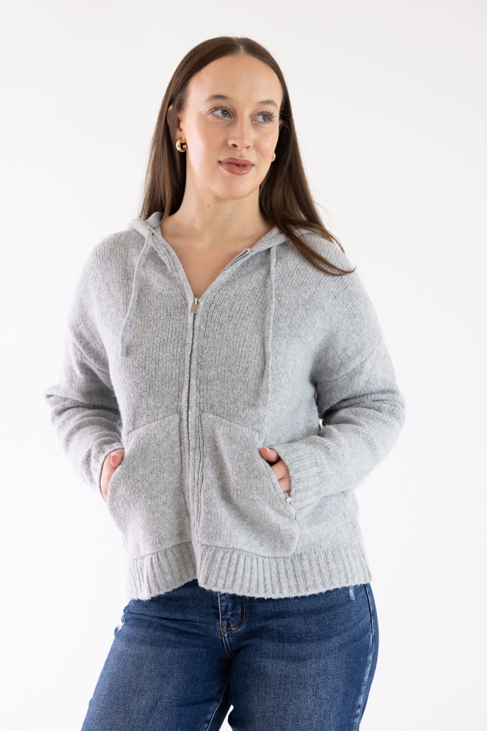 A woman wearing a light gray zip-up hoodie with drawstrings and front pockets, paired with blue jeans, standing against a plain white studio background.