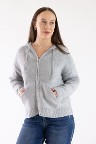 A woman wearing a light gray zip-up hoodie with drawstrings and front pockets, paired with blue jeans, standing against a plain white studio background.