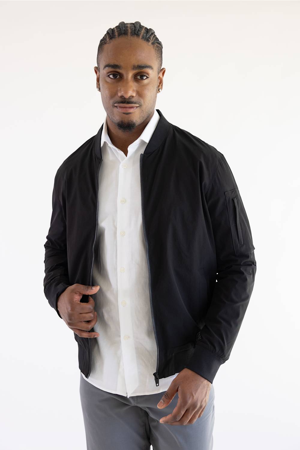 A man wearing a black zip-up bomber jacket over a white button-down shirt and gray trousers, standing against a plain white studio background.