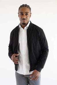 A man wearing a black zip-up bomber jacket over a white button-down shirt and gray trousers, standing against a plain white studio background.