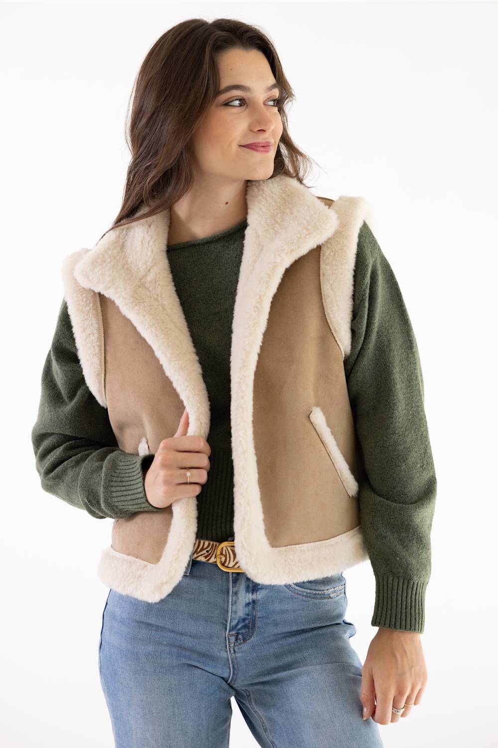 A woman wearing a tan shearling-lined sleeveless vest over an olive green knit sweater and blue jeans, photographed against a clean white studio background.