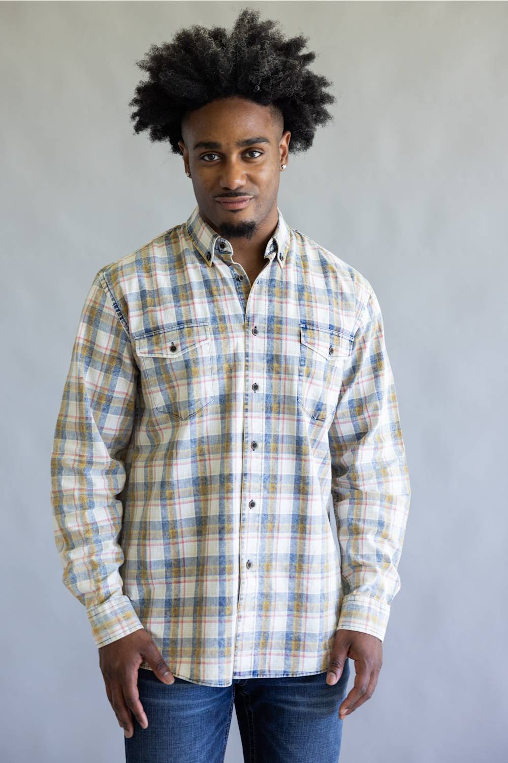 A man wearing a long-sleeve button-down plaid shirt in beige, blue, and white, paired with blue jeans, standing against a plain gray studio background.
