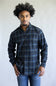 A man wearing a dark blue and black plaid button-down shirt with blue jeans, standing against a plain light gray background.