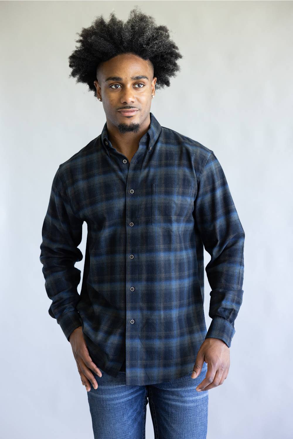 A man wearing a dark blue and black plaid button-down shirt with blue jeans, standing against a plain light gray background.