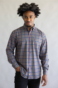 A man with dark curly hair wearing a long-sleeve button-down plaid shirt in blue, red, and gray, paired with black pants, standing against a light gray background.