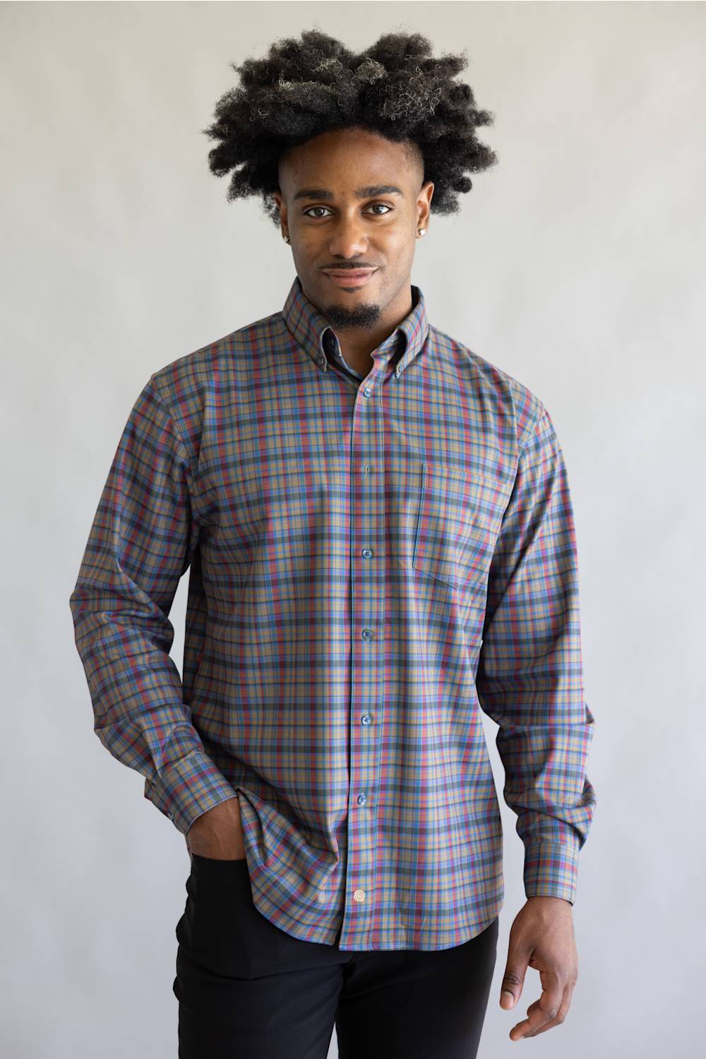 A man with dark curly hair wearing a long-sleeve button-down plaid shirt in blue, red, and gray, paired with black pants, standing against a light gray background.