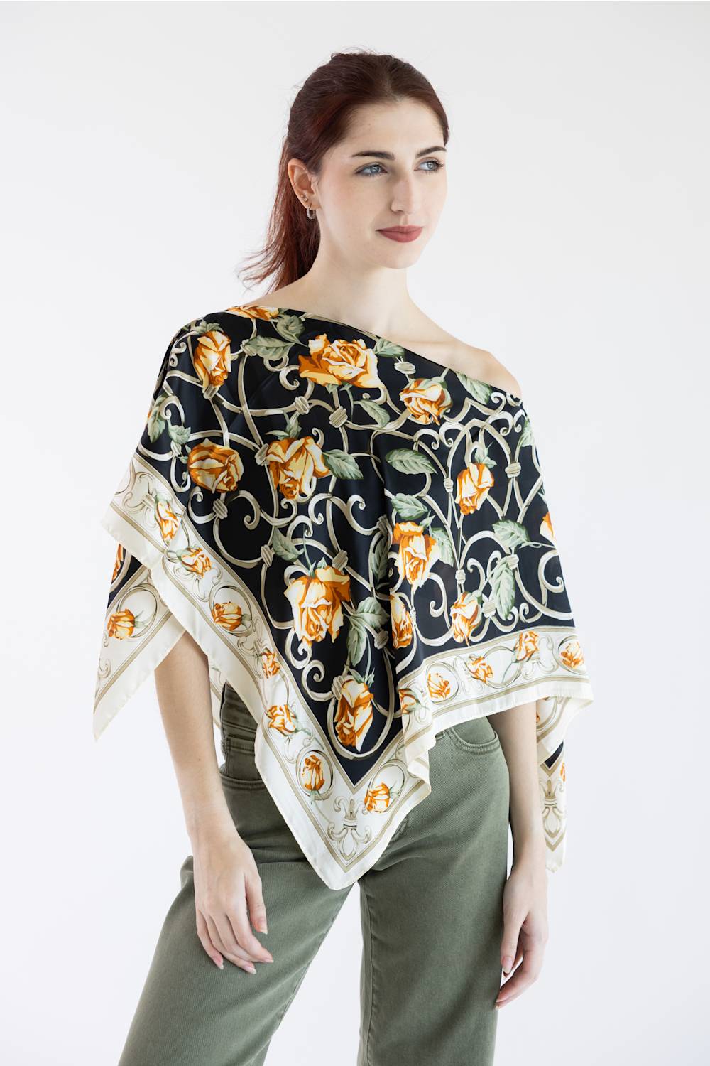 A woman modeling a square scarf top draped off her shoulders, featuring a black background with a yellow and orange floral pattern and a cream border, paired with olive green pants against a white studio background.