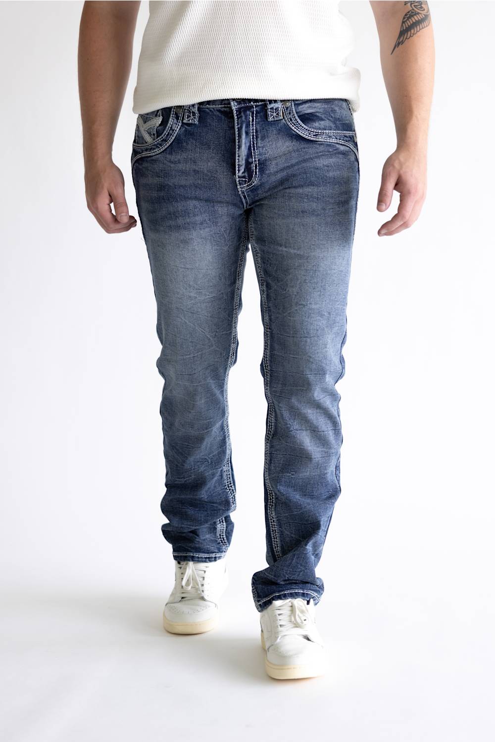 A person wearing medium-wash blue jeans with a faded wash, paired with a white t-shirt and white sneakers, shown from the waist down against a white studio background.