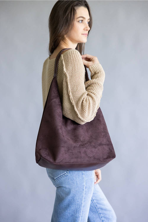 A woman wearing a beige knit sweater and light blue jeans carries a large dark brown suede shoulder bag against a plain gray studio background.