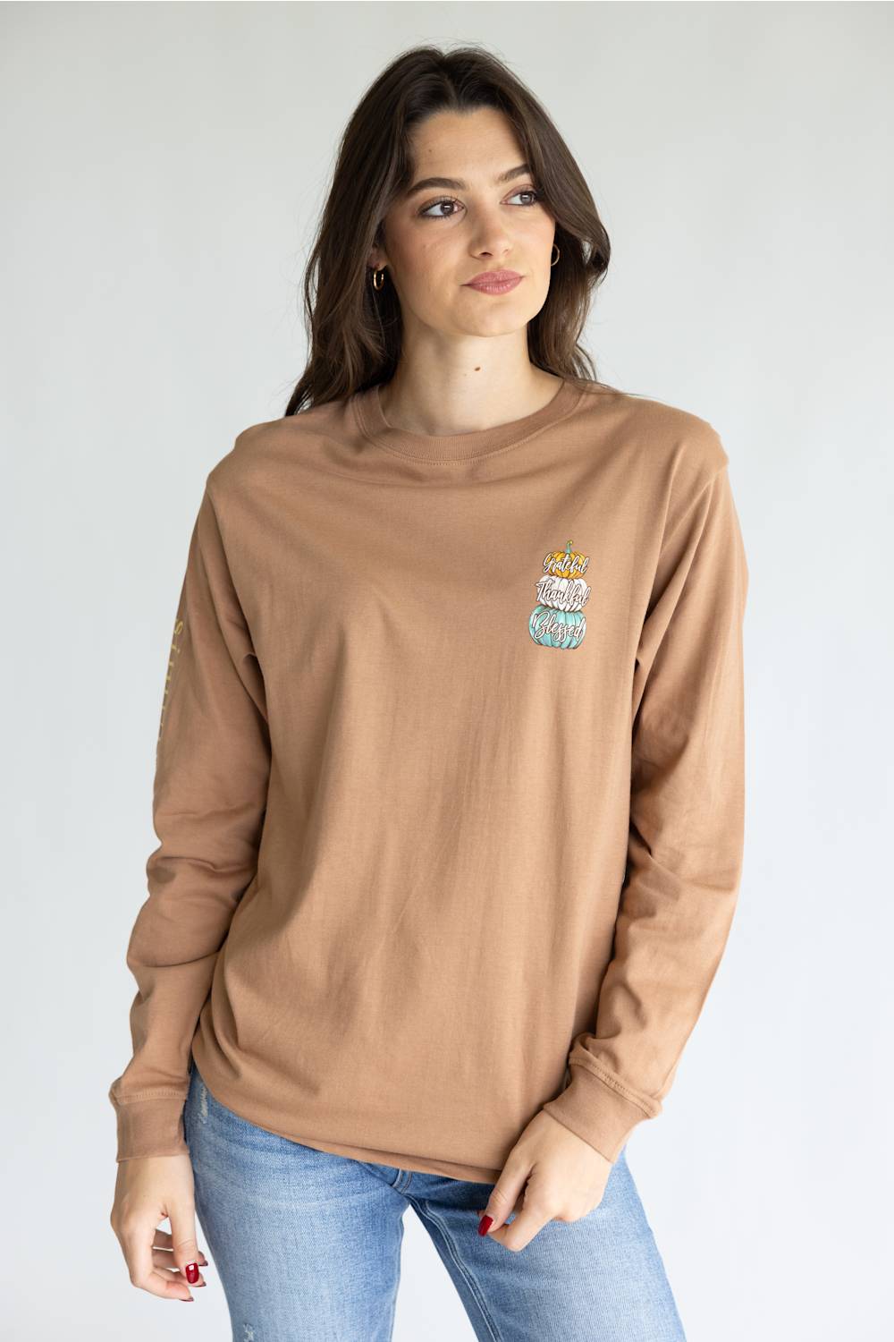 A woman modeling a tan long-sleeve crewneck sweatshirt with a small embroidered graphic on the left chest, paired with blue jeans, against a plain light gray background.