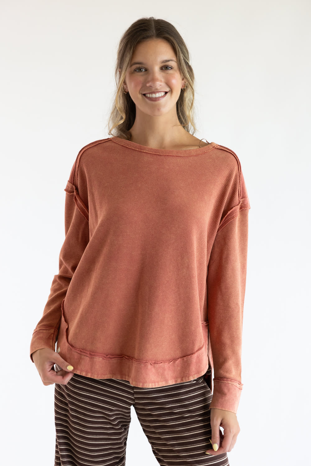 A woman modeling a dusty rose long-sleeve knit top with a wide boat neckline and relaxed fit, paired with brown and white striped pants, against a plain white studio background.