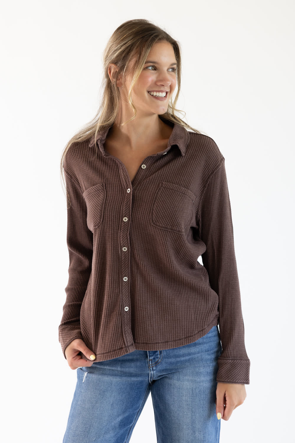A woman wearing a brown ribbed long-sleeve button-up shirt and blue jeans, standing against a plain white studio background.
