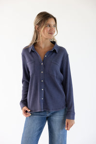 A woman wearing a dark blue ribbed long-sleeve button-up shirt and blue jeans, standing against a plain white background.