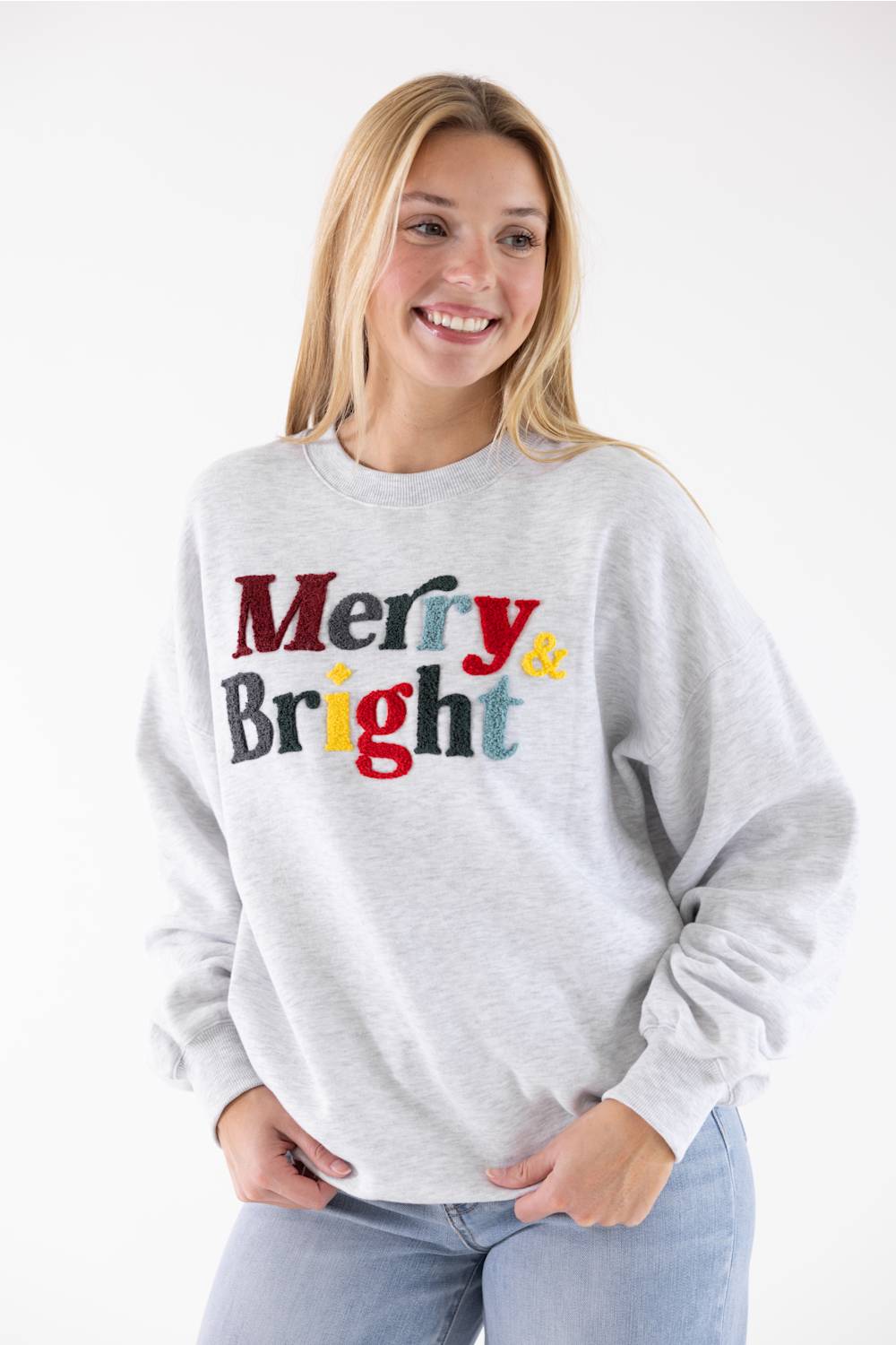A person wearing a light heather gray crewneck sweatshirt with the words 'Merry & Bright' printed across the chest in multicolored textured letters.