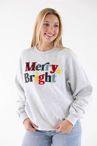 A person wearing a light heather gray crewneck sweatshirt with the words 'Merry & Bright' printed across the chest in multicolored textured letters.