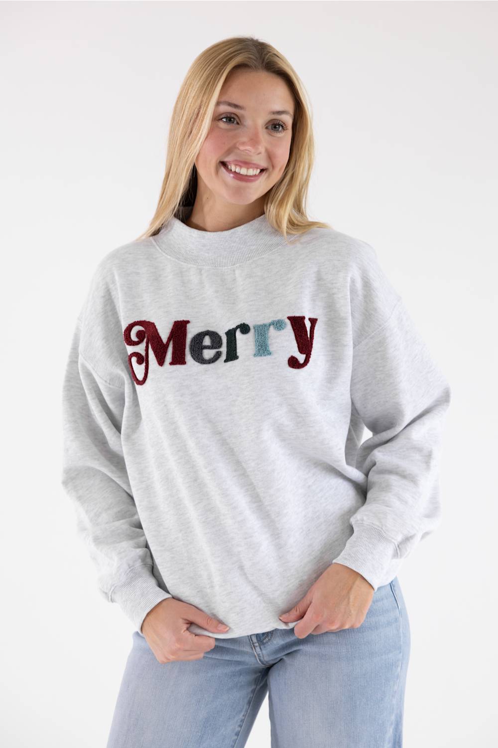 A smiling blonde woman wearing a light gray crewneck sweatshirt with 'Merry' written across the chest in maroon and blue glitter-style letters, paired with light blue jeans, against a plain white studio background.