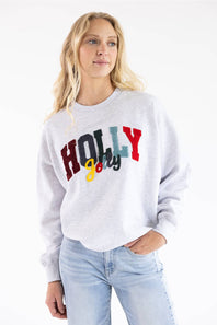 A woman wearing a light gray heather crewneck sweatshirt with a colorful 'HOLLY jolly' graphic on the front, paired with light blue jeans, against a plain white background.