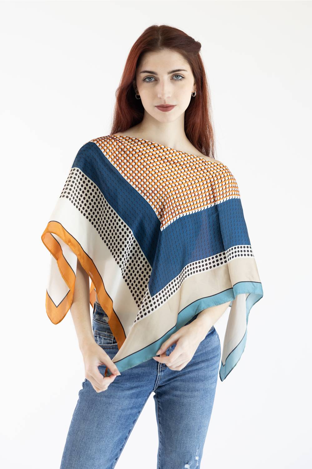 A woman with red hair wearing a multi-colored patterned poncho top and blue jeans, standing against a plain white background.