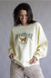 A woman wearing a cream-colored crewneck sweatshirt with a 'Club Retriever Stay Golden' graphic featuring a golden retriever, paired with beige corduroy pants, against a neutral gray studio background.