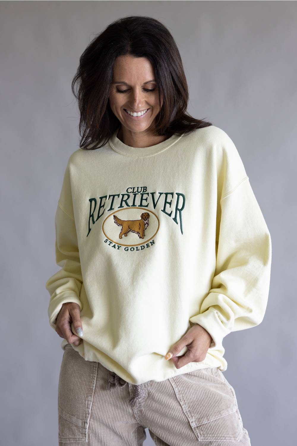 A woman wearing a cream-colored crewneck sweatshirt with a 'Club Retriever Stay Golden' graphic featuring a golden retriever, paired with beige corduroy pants, against a neutral gray studio background.