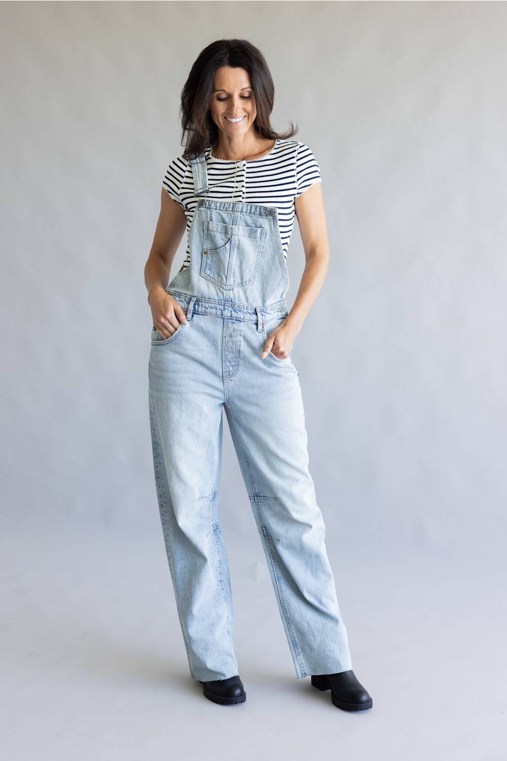 A woman modeling light-wash denim overalls over a black and white striped t-shirt, standing against a neutral gray studio background.