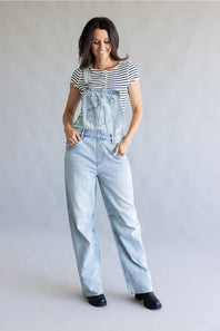 A woman modeling light-wash denim overalls over a black and white striped t-shirt, standing against a neutral gray studio background.