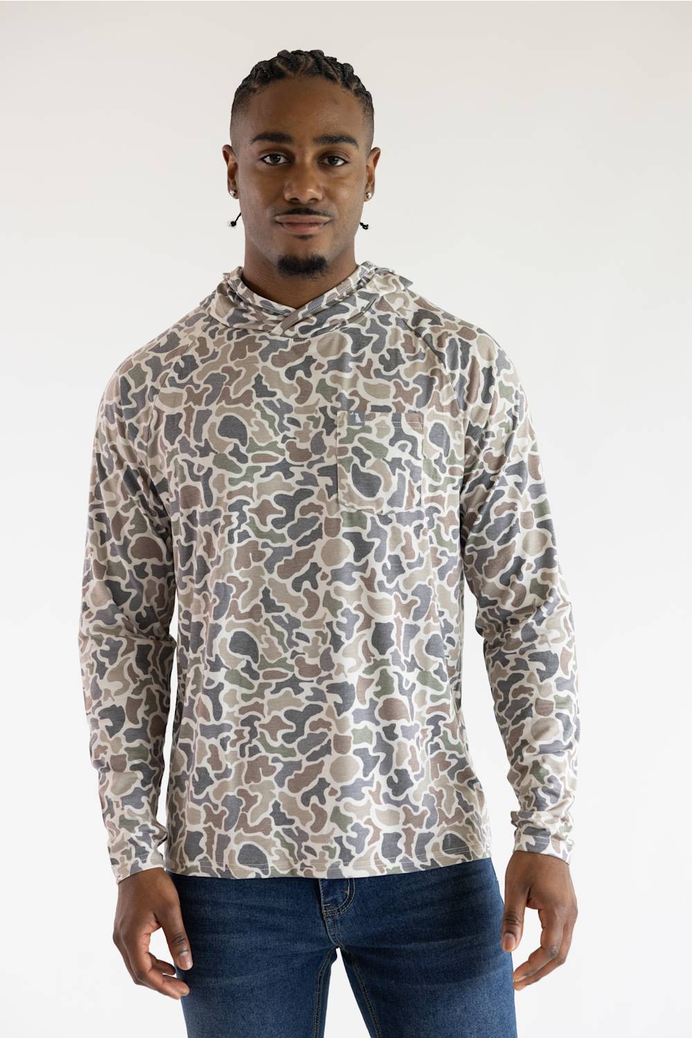 A man wearing a long-sleeve hooded shirt with an abstract organic camouflage pattern in beige, brown, and gray, paired with blue jeans, against a plain white studio background.
