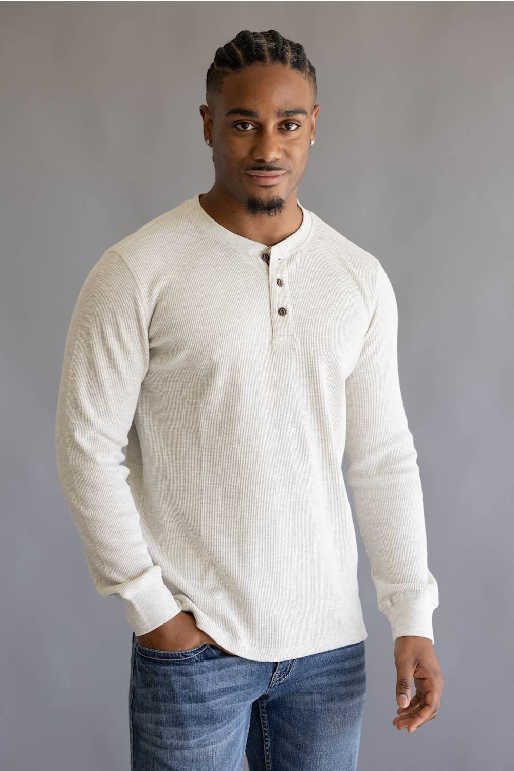 A man wearing a light cream-colored long-sleeve henley shirt and blue jeans, standing against a solid gray studio background.
