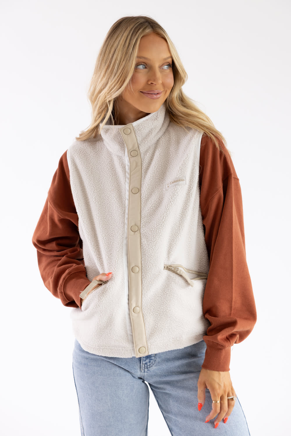 A woman wearing a two-tone fleece jacket with a cream body and rust-colored sleeves, paired with light blue jeans, against a plain white background.
