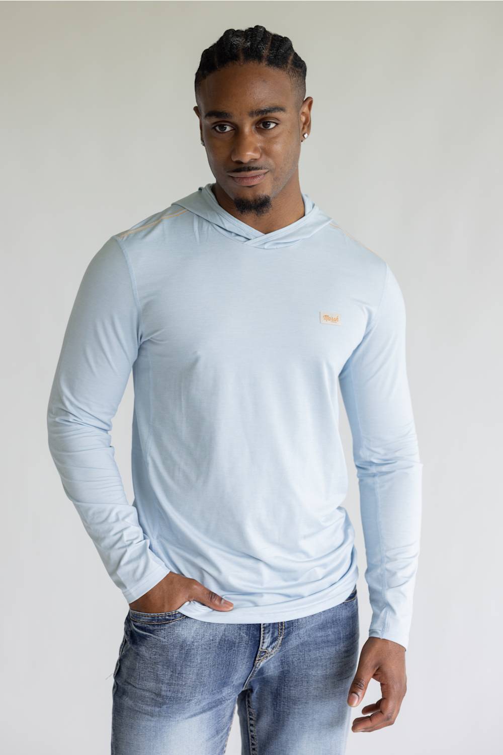 A man wearing a light blue long-sleeve hooded shirt and blue jeans, standing against a plain light gray background.