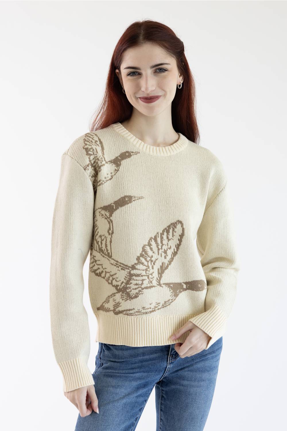 A woman with red hair wearing a cream-colored knit sweater with a brown graphic print of flying ducks, paired with blue jeans, against a plain white background.