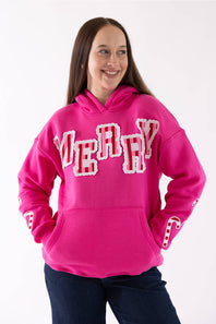 A woman wearing a bright pink pullover hoodie with 'MERRY' written across the chest in large white letters outlined in red, paired with dark blue jeans, against a plain white background.