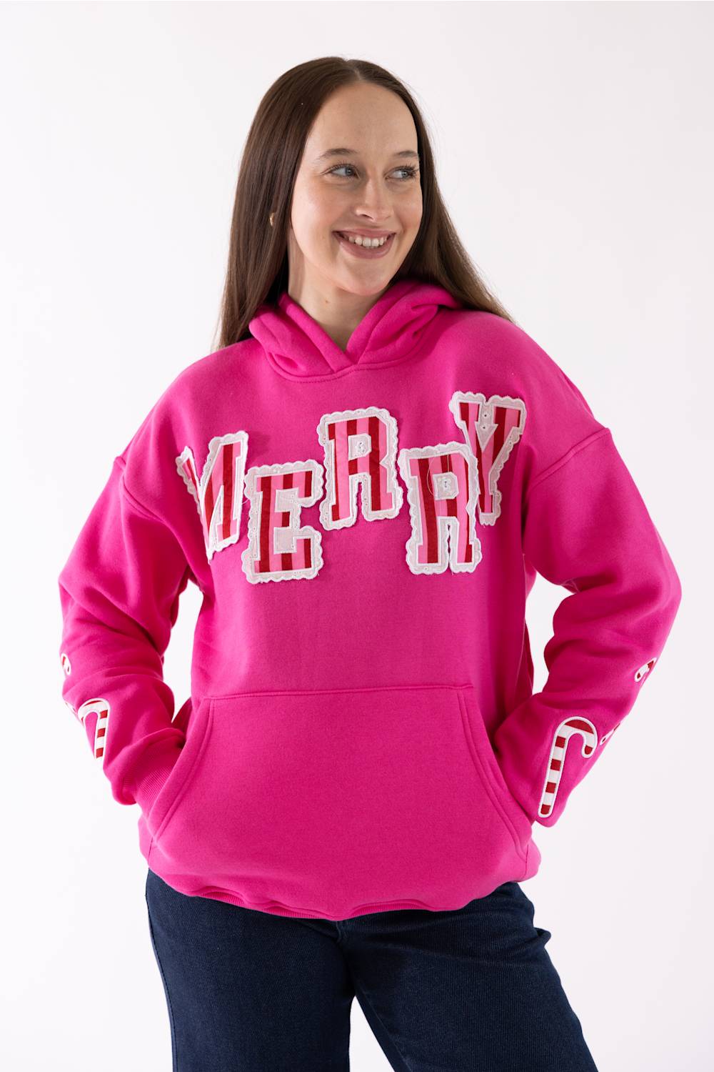 A woman wearing a bright pink pullover hoodie with 'MERRY' written across the chest in large white letters outlined in red, paired with dark blue jeans, against a plain white background.