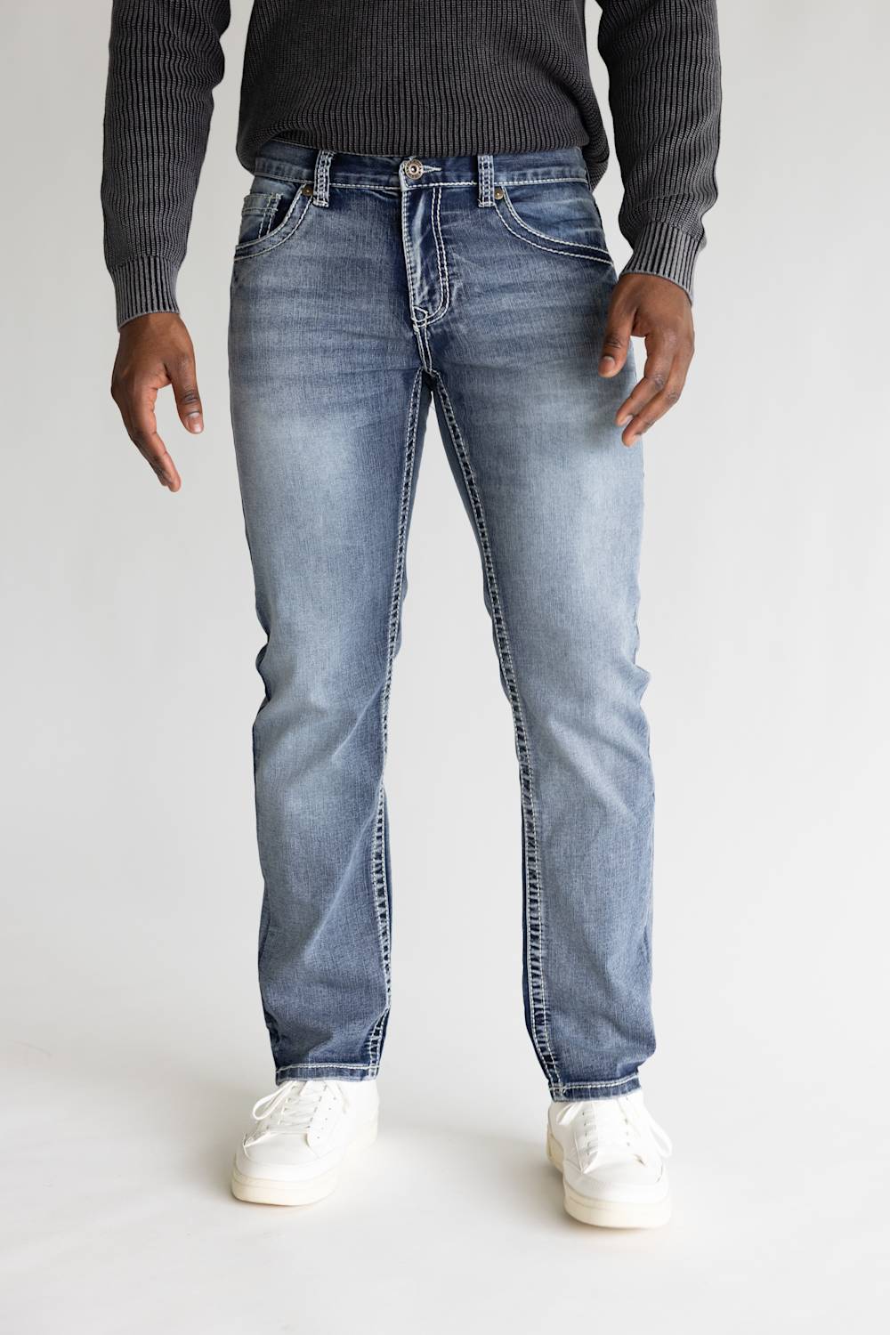 A person wearing light-wash blue jeans with a dark ribbed sweater and white sneakers, standing against a plain light gray studio background.