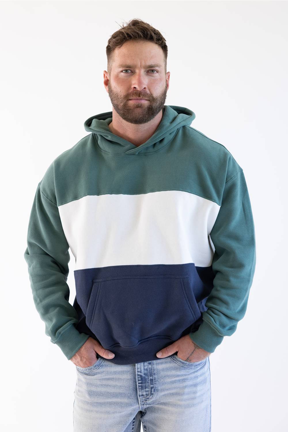 A man wearing a color-blocked hoodie with a dark green upper section, a wide white middle stripe, and a navy blue lower section, paired with light-wash jeans against a plain white background.